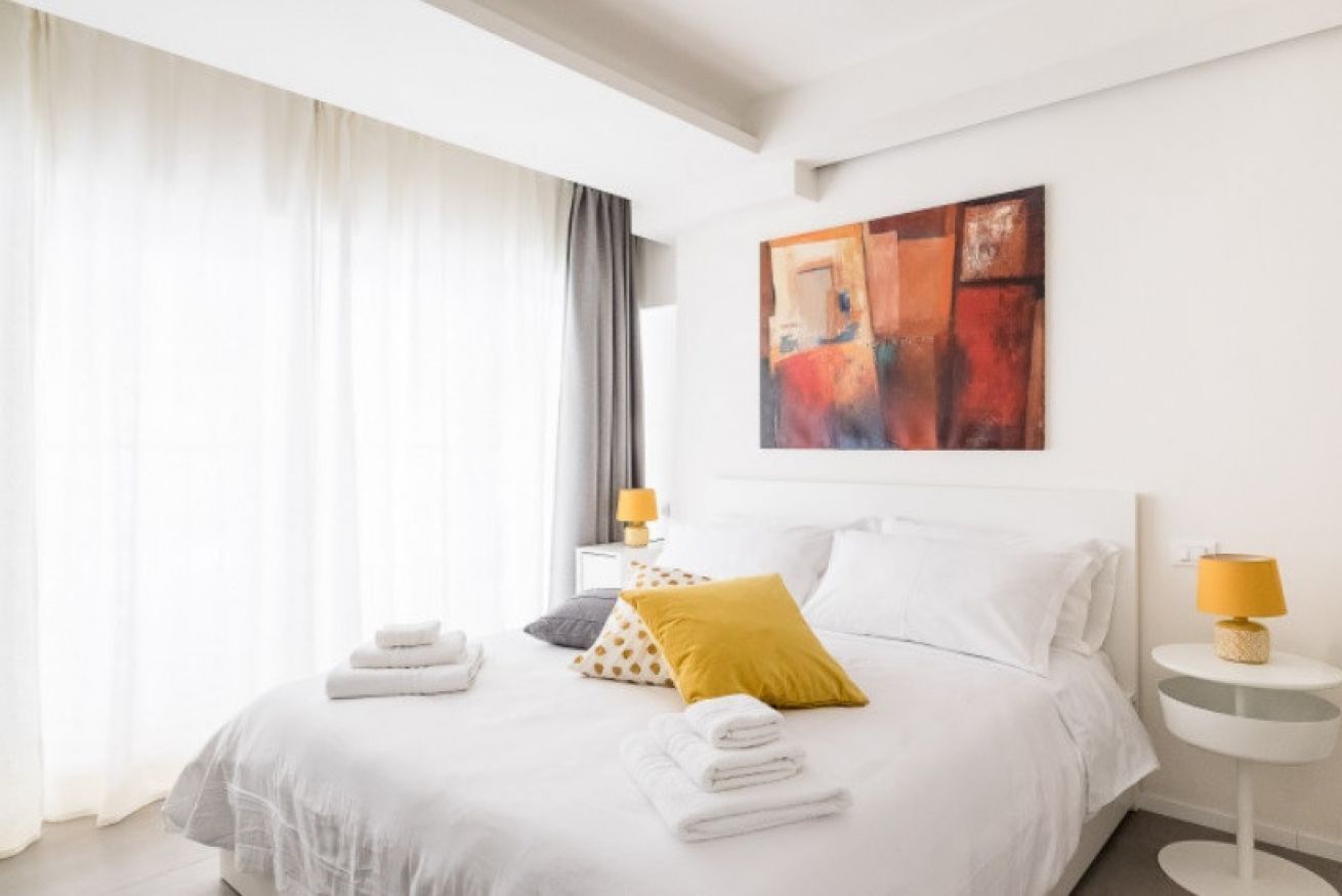 A modern hotel room with a neatly made bed, a large abstract painting on the wall, and a side table with a lamp.
