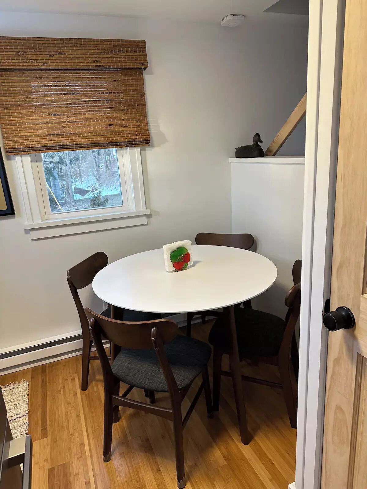 A dining area with a round table and four chairs, a window with blinds, and a view of trees outside.