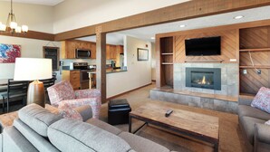 A spacious living room with a fireplace, a television mounted on the wall, and a kitchen area in the background. - Three Bedroom Townhouse. AMAZING VIEWS. On bus route. Pet friendly. (Breckenridge)