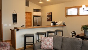 Een moderne keuken met een bar, houten kasten en een koelkast. Er is een grijze bank met een kussen met patroon en een eettafel met stoelen.