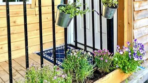 Una casa di legno con una porta bianca, una piccola finestra e un portico con ringhiere nere. Ci sono piante in vaso appese alle ringhiere e un tappetino all'ingresso sul portico.