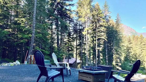 Zona salotto all'aperto con poltrone Adirondack e un braciere, circondata da alberi alti e una montagna sullo sfondo.