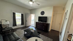 A living room with a black leather sofa, a coffee table with a plant, a TV on a wooden cabinet, and a ceiling fan.