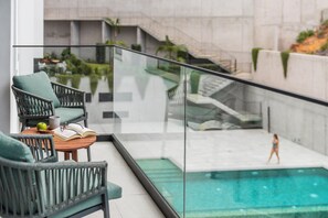 A balcony with a glass railing, a wicker chair, a small table with a book and an apple, and a view of a swimming pool and a person walking. - Funchal, with pool - Uptown 13 apartment (Funchal)