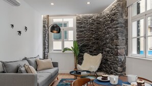 A modern living room with a grey sofa, a dining table with chairs, a stone accent wall, and a large window with shutters.