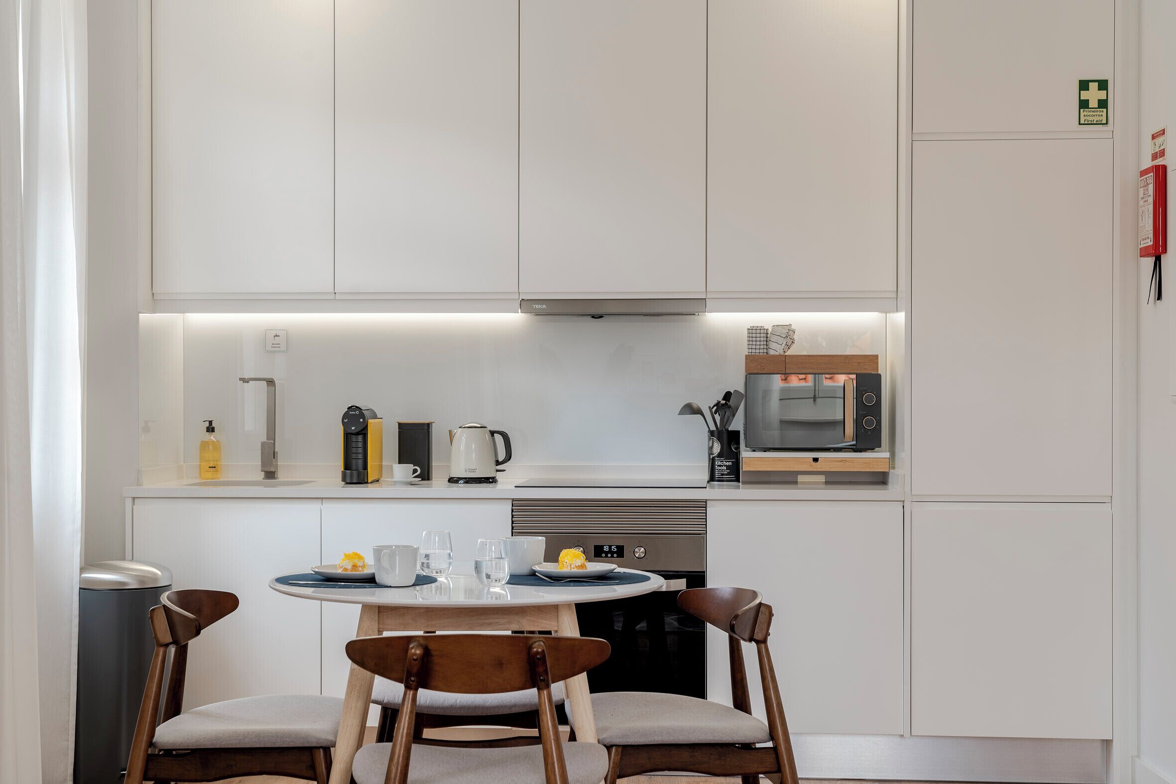Une cuisine moderne avec des meubles blancs, un four à micro-ondes, une bouilloire et un lave-vaisselle. On y trouve une table ronde avec des chaises et un petit panier à fruits.