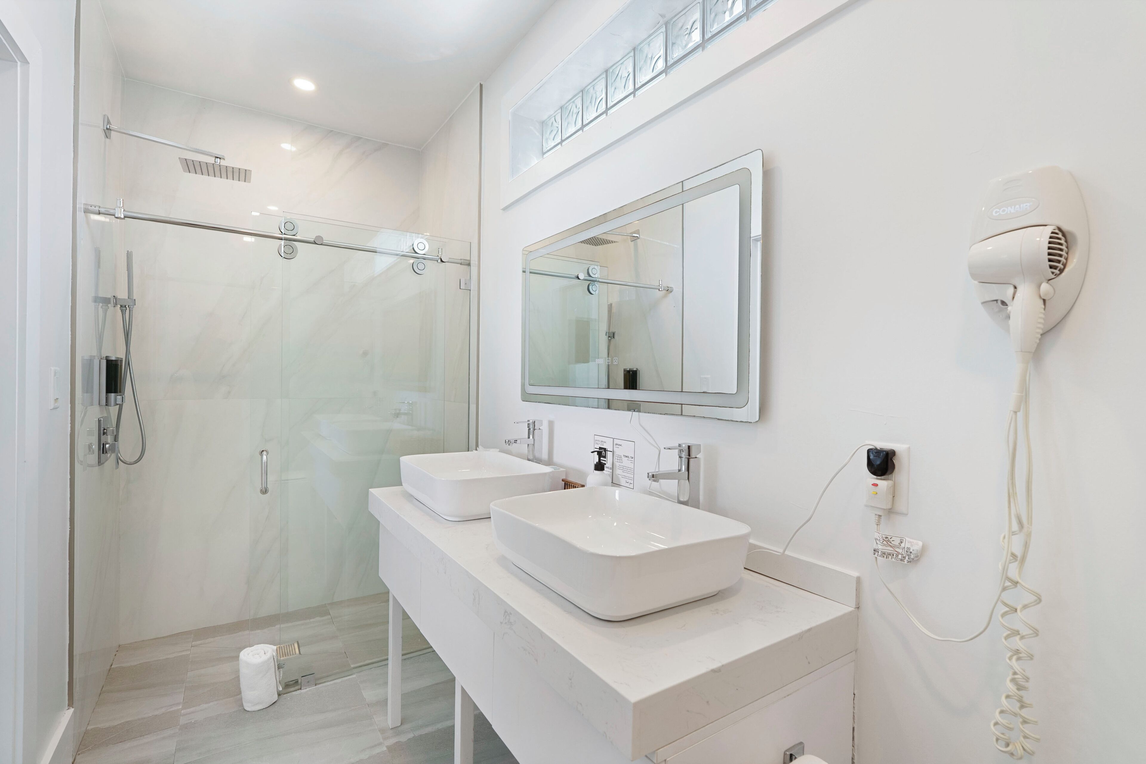 A modern bathroom with two sinks, a glass shower enclosure, a hairdryer, and a mirror.