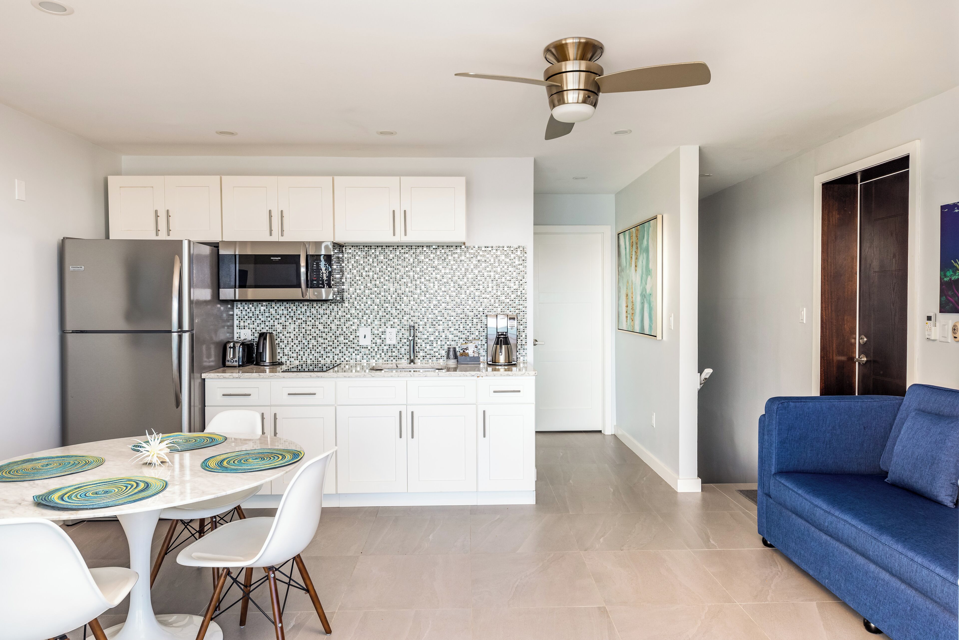 Una cocina moderna con armarios blancos, un refrigerador de acero inoxidable y un microondas. Hay una mesa de comedor con sillas blancas y un sofá azul en la sala.