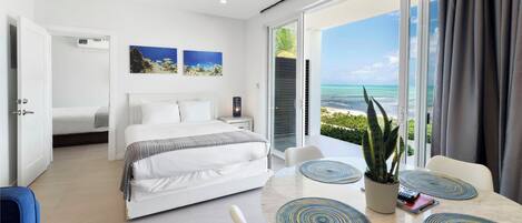 A modern hotel room with a bed, a dining area with a table and chairs, a ceiling fan, and a view of the ocean through large windows.