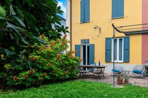 A yellow building with blue shutters, a patio with a table and chairs, and a large bush with orange flowers.