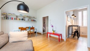 A modern living room with a sofa, a dining table with chairs, a bookshelf, a red bench, and a black floor lamp.