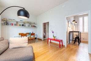 Ein modernes Wohnzimmer mit einer Couch, einem Esstisch mit Stühlen, einem Bücherregal, einer roten Bank und einer schwarzen Stehlampe.