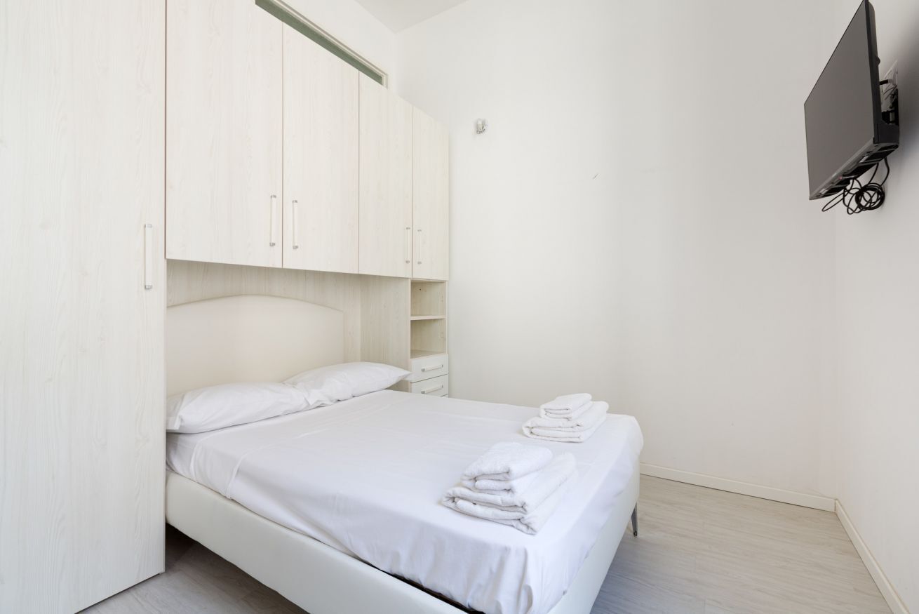 A bedroom with a bed, white curtains, a flat-screen TV mounted on the wall, and a wooden wardrobe.