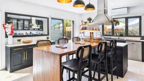 A modern kitchen with a central island, black chairs, pendant lighting, and a large window offering a view of the outdoors. - Stunning!! - Casa Chameleon - Sleeps 14 - Close to Quinta Do Lago & Vale Do Lobo (Faro)