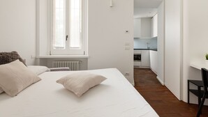 A bedroom with a large bed, a window, a radiator, and a view into a kitchen area. - GuestHost - Nice and small Studio-Milan Central Station (Milano)