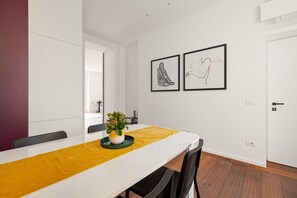 A modern dining area with a white table, yellow table runner, and black chairs. There are two framed artworks on the wall and a potted plant on the table.