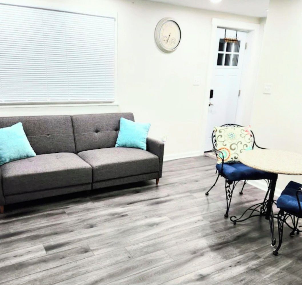 A room with a grey sofa, a dining table with chairs, a clock on the wall, and a door in the background.