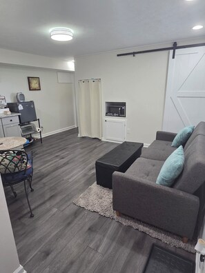 A modern living room with a grey sofa, a coffee table, a dining area with a small table and chairs, a microwave, and a door with a decorative handle.