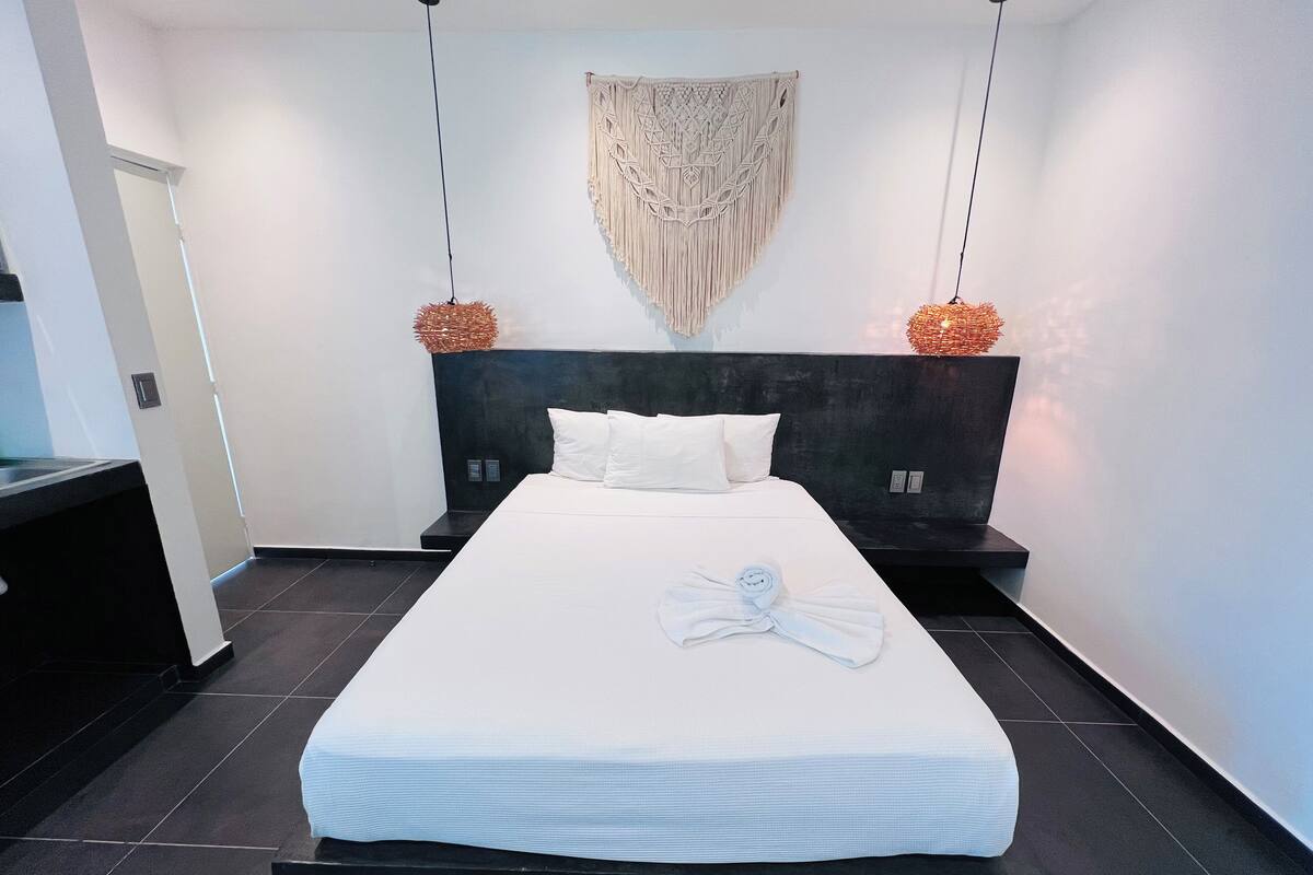 A modern hotel room with a large bed, a headboard, and a decorative macrame wall hanging.