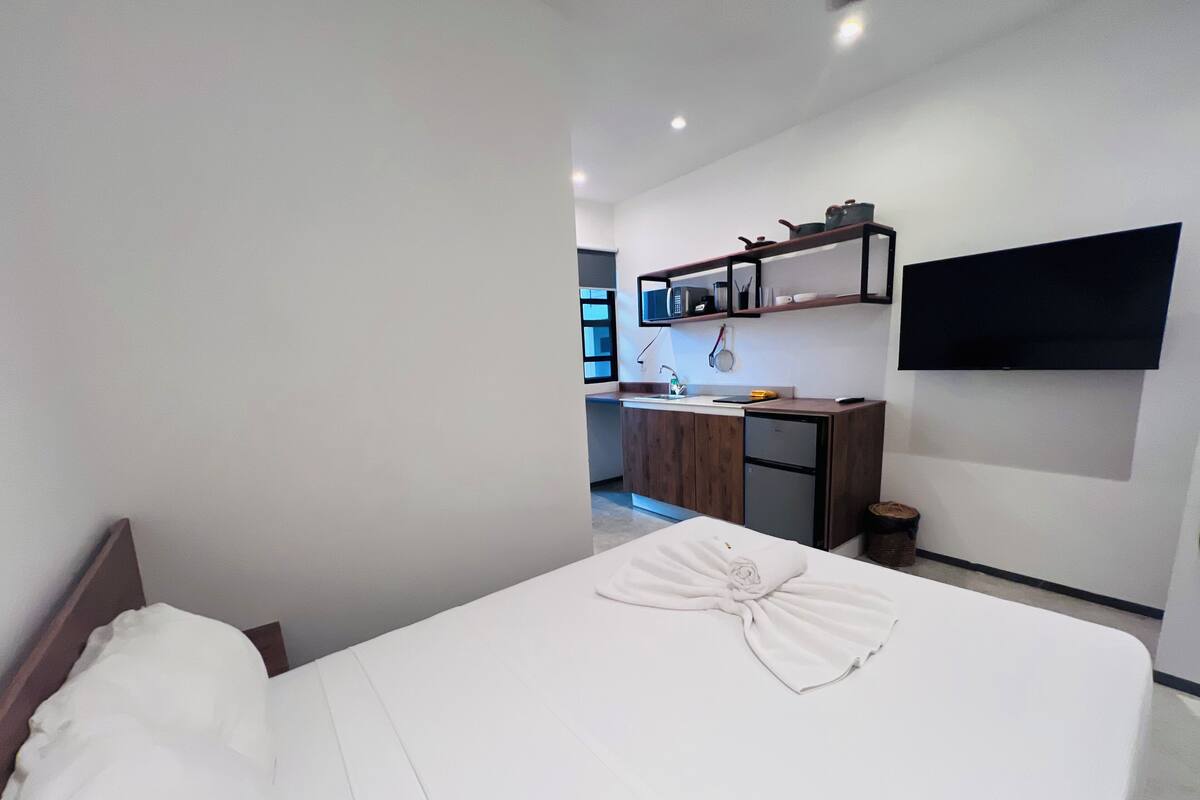 A hotel room with a neatly made bed, a small kitchenette with a refrigerator, and a flat-screen TV mounted on the wall.