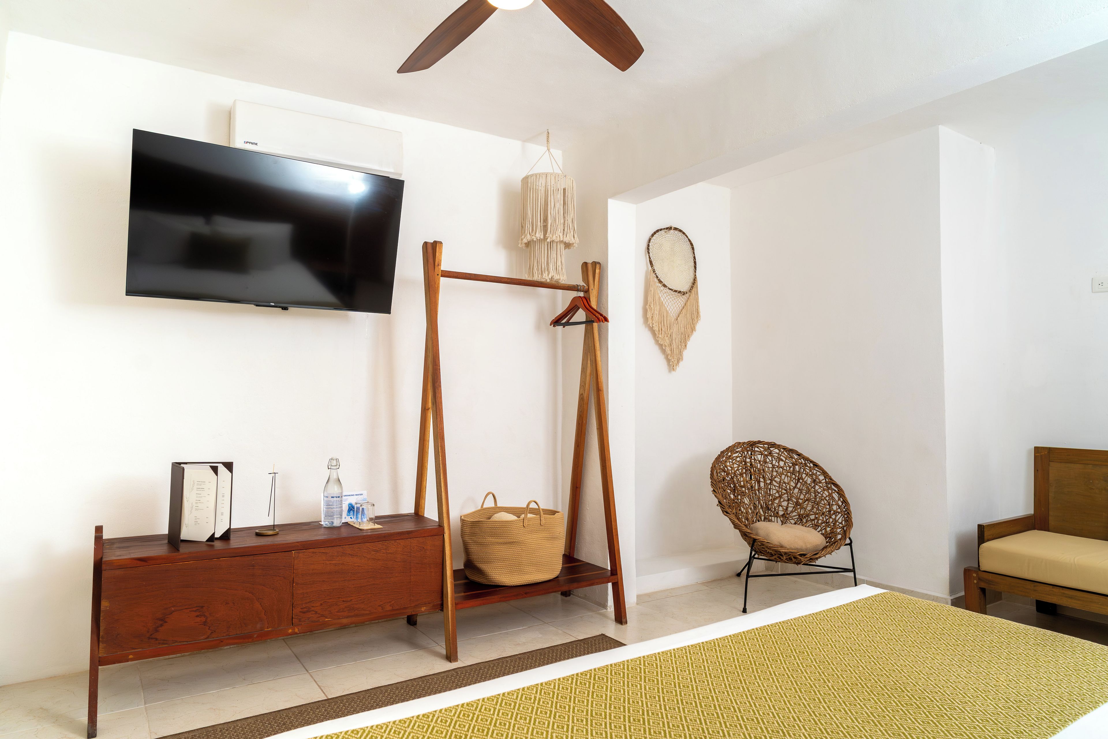 Een kamer met een houten dressoir, een aan de muur bevestigde televisie, een macramé decoratie, een rieten stoel en een bed met een geel geboeide vloerkleden.