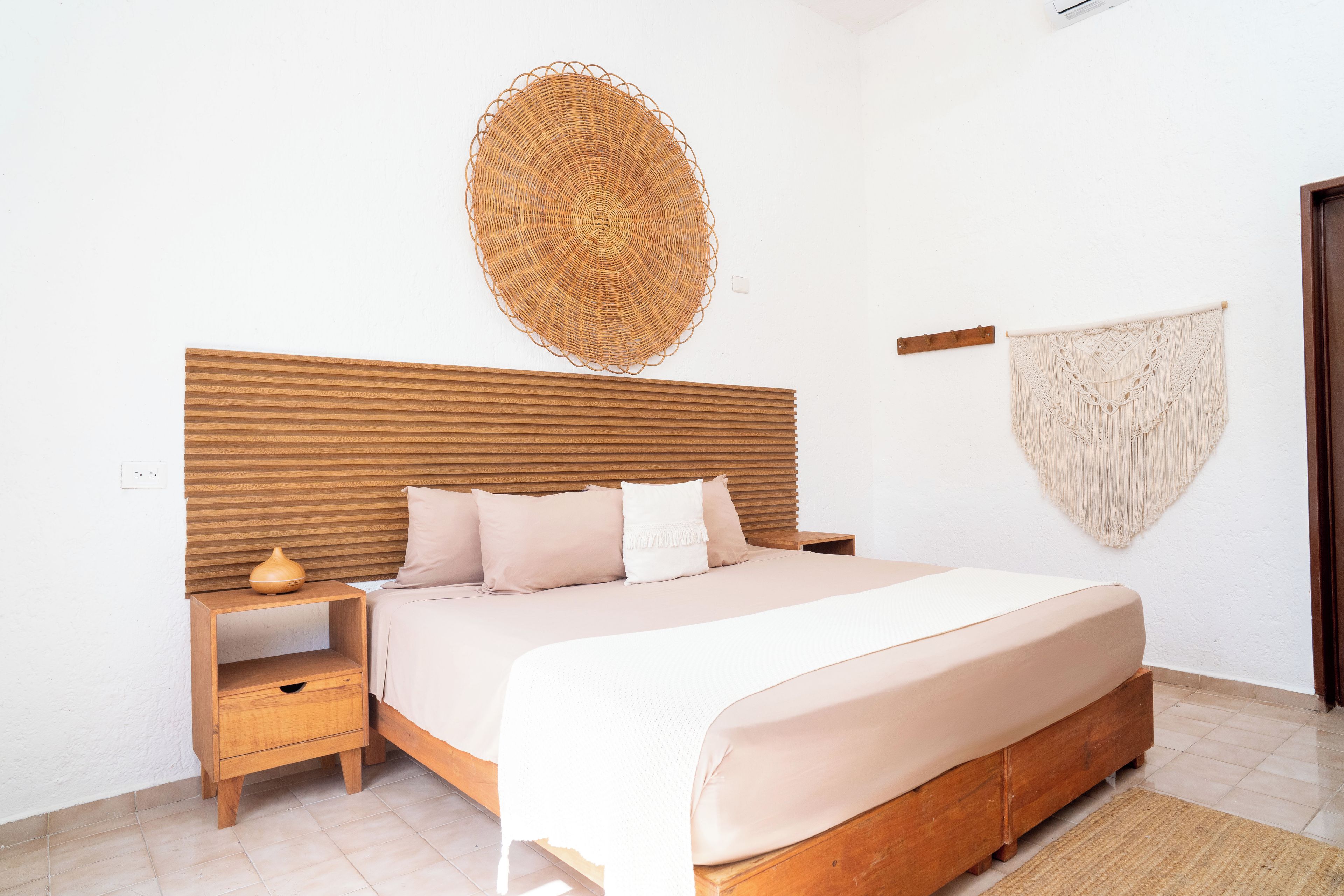 A bedroom with a wooden headboard, a bed with a beige blanket, a woven wall hanging, and a macrame wall hanging.