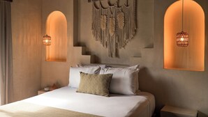 Une chambre avec un lit, des oreillers, une décoration murale en macramé et deux lampes suspendues.