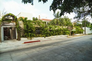 Vista de la calle con un edificio que tiene un acceso cubierto, una acera roja y un paseo marítimo con plantas en macetas.