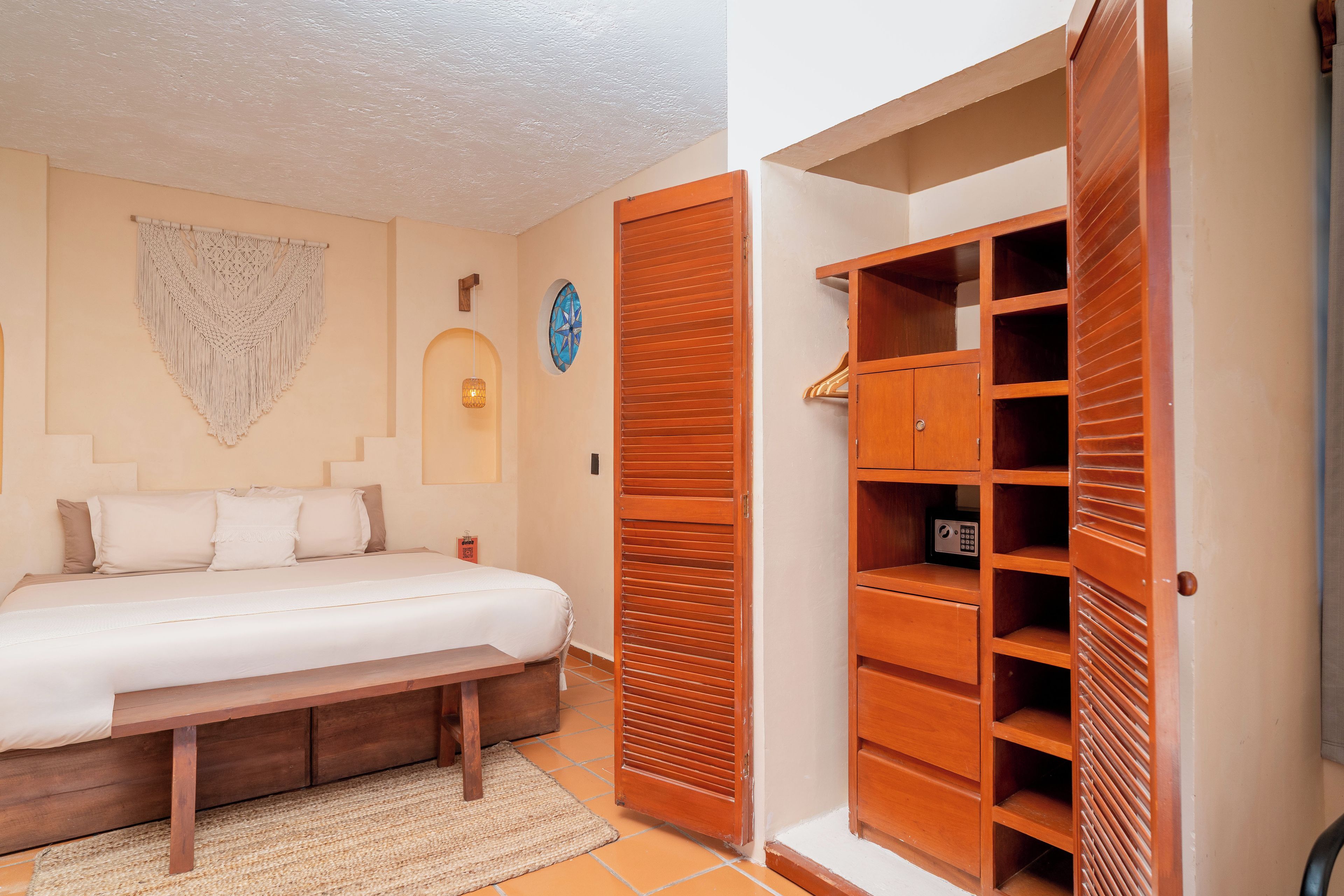 A bedroom with a bed, a wooden wardrobe, a macrame wall hanging, and a wall-mounted light fixture.