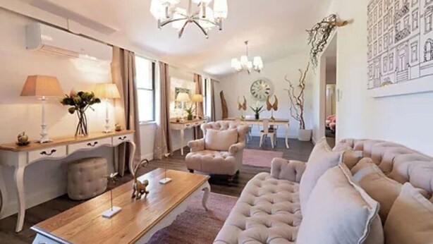 Amplia sala de estar con un sofá grande color beige, una mesa de centro de madera y una araña. Hay un comedor con mesa y sillas, y una cocina al fondo.