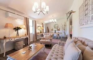 A spacious living room with a large beige sofa, a wooden coffee table, and a chandelier. There is a dining area with a table and chairs, and a kitchen in the background. - King Apartment One (Aldgate)