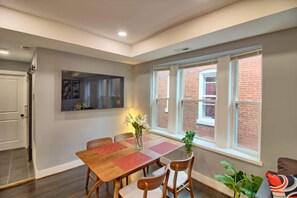 Un comedor con una mesa y sillas de madera, un florero con flores y una ventana con vista a edificios de ladrillo.