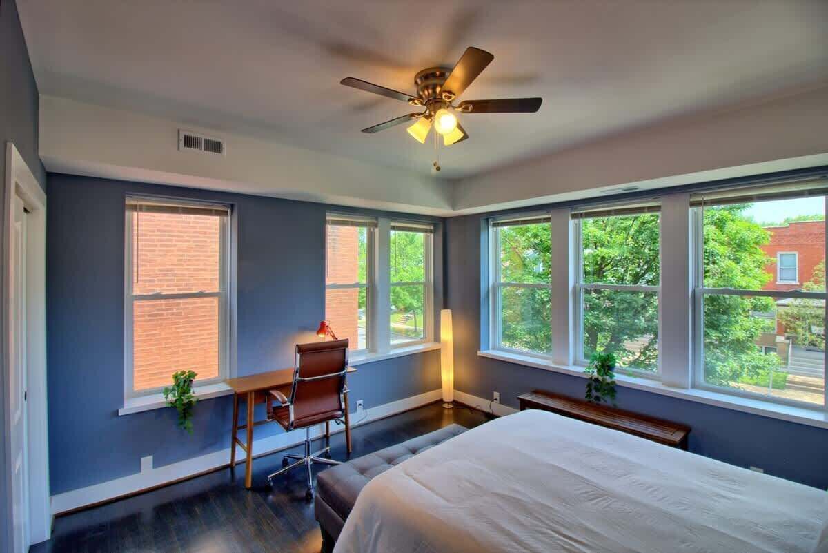 A bedroom with a bed, a desk, a chair, a ceiling fan, and large windows.