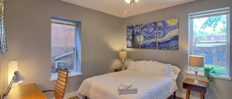 A bedroom with a bed, a desk, a chair, a ceiling fan, and artwork on the wall.