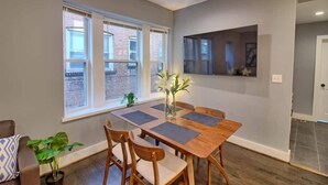 Un comedor con una mesa y sillas de madera, un jarrón con flores y una ventana con vista a un edificio de ladrillo.