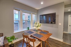 Un comedor con una mesa y sillas de madera, un jarrón con flores y una ventana con vista a un edificio de ladrillo.