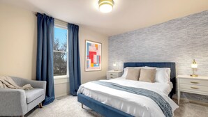 A bedroom with a large bed, a grey armchair, a window with blue curtains, a wall art piece, and a textured wall.