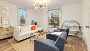 A modern living room with a white sofa, a glass coffee table, and a blue armchair. There is a wooden sideboard with drawers, a round white table, and a decorative ceiling light.