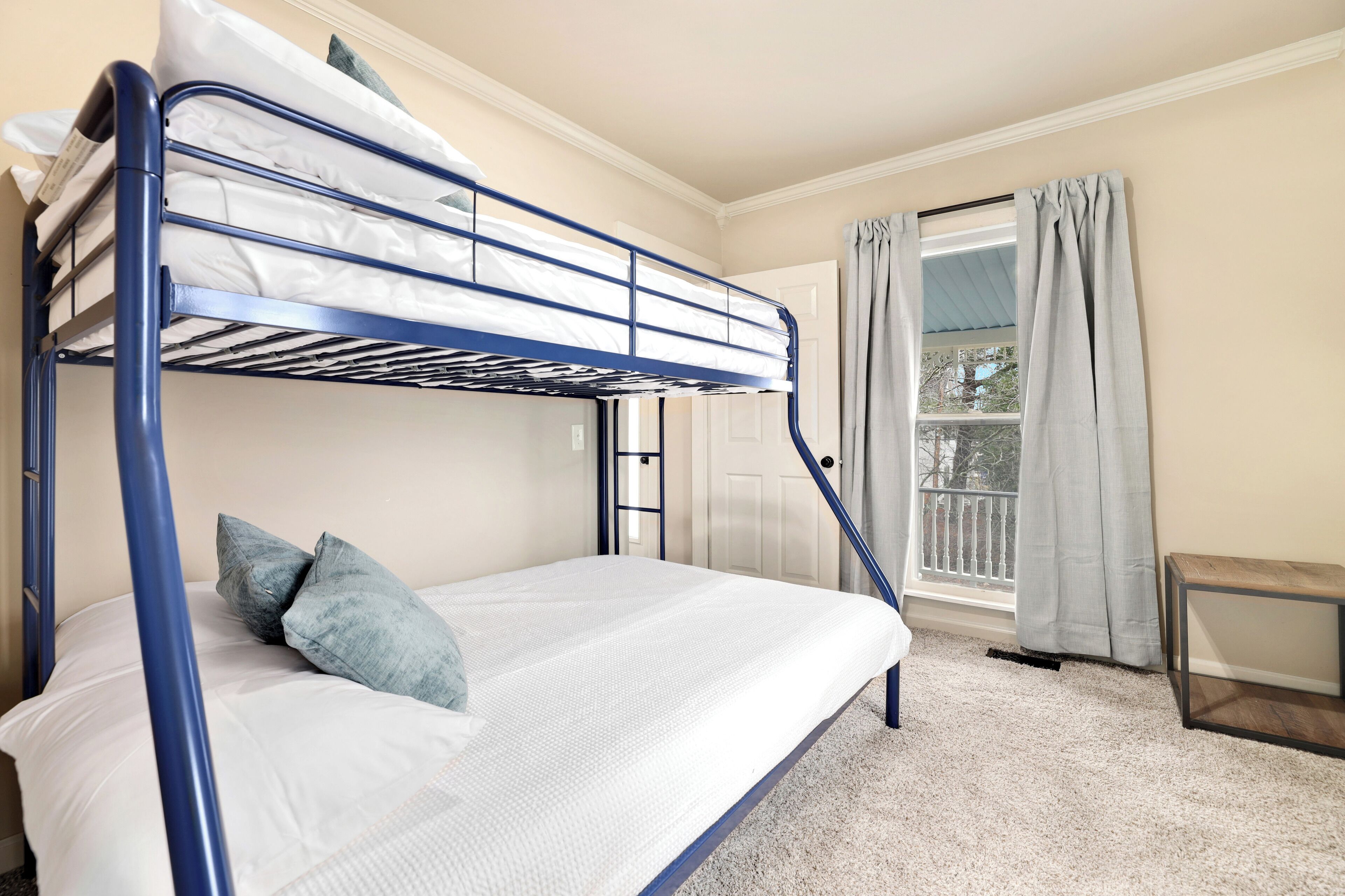 A bunk bed with a white mattress and blue metal frame in a room with beige walls, a window with curtains, and a small wooden bench.