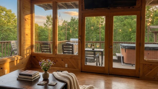 Una cabaña de madera con televisor, una mesa con libros y un florero, y vistas a una terraza con asientos exteriores y un jacuzzi.