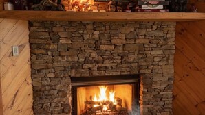 Una chimenea de piedra con fuego, un estante superior con libros y objetos decorativos, y una manta acogedora en el suelo.