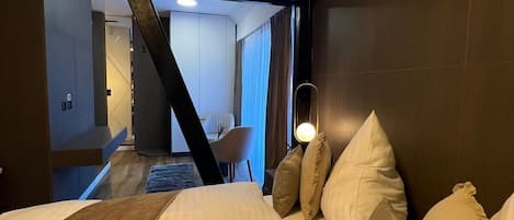A hotel room with a large bed, a desk, and a chair. There is a view of another room in the background.