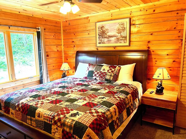 A wooden cabin bedroom with a large bed, a headboard, two bedside tables, a lamp, and a painting on the wall.