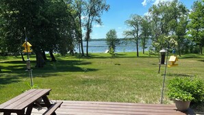 Una terraza de madera con mesas de picnic, rodeada de césped y árboles, con vista a un lago.