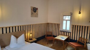 A corner room with a bed, a table, two chairs, a window, and a wall-mounted light fixture.