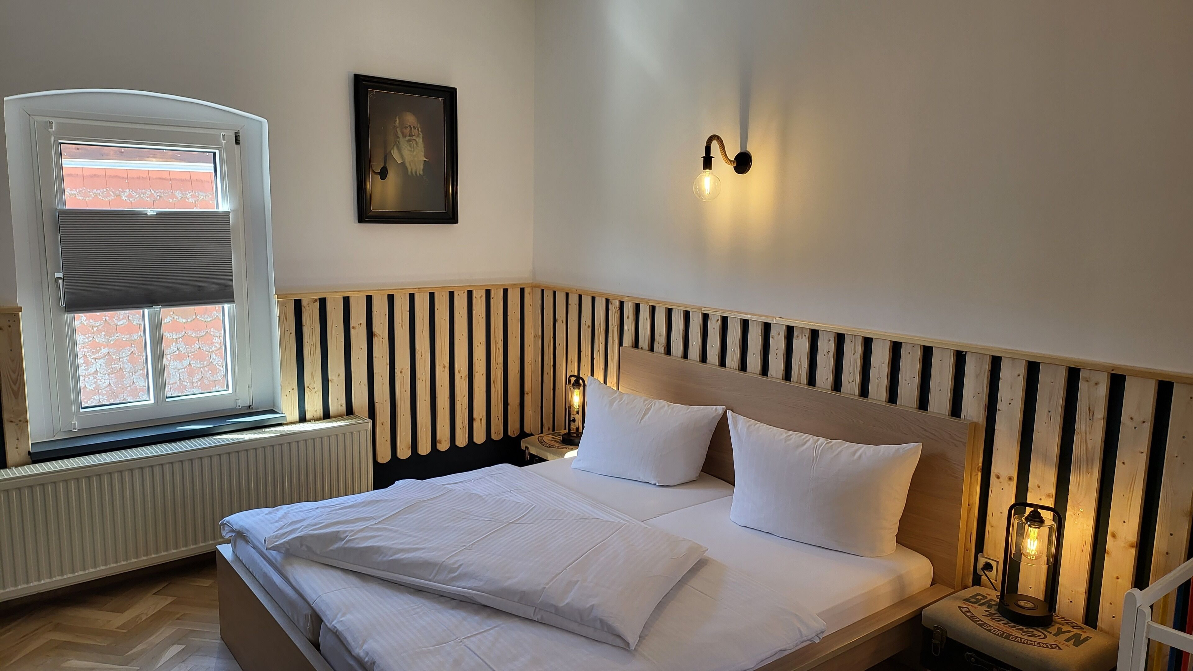 A bedroom with a wooden headboard, a large bed with white bedding, a window with blinds, a wall-mounted light fixture, and a small table with a lamp.