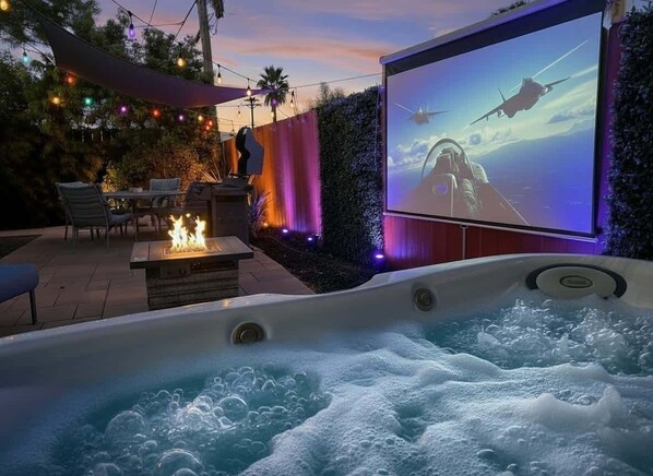 Un jacuzzi con una gran pantalla mostrando una escena de película, rodeado de asientos exteriores y luces colgantes.