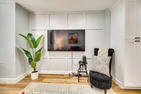 A modern living room with a flat-screen TV mounted on a white wall, a large potted plant, a black velvet armchair, and a patterned rug.