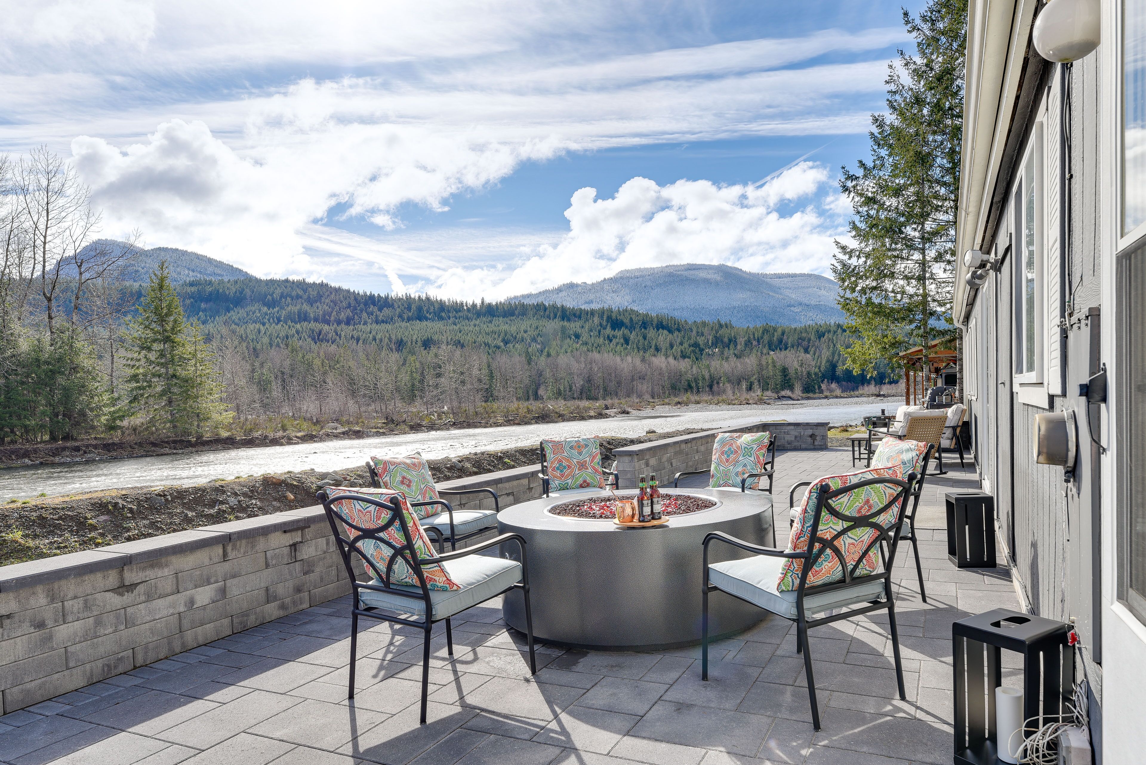 An outdoor patio with a circular fire pit, seating, and a view of a river and mountains.