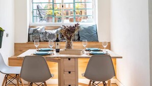 A dining area with a wooden table set for two, two chairs, a vase with dried flowers, a bottle of wine, and a window with a view of the outside. - BBQ, Easy Parking & near Tejo, Lisbon Garden Stays (Lisboa)